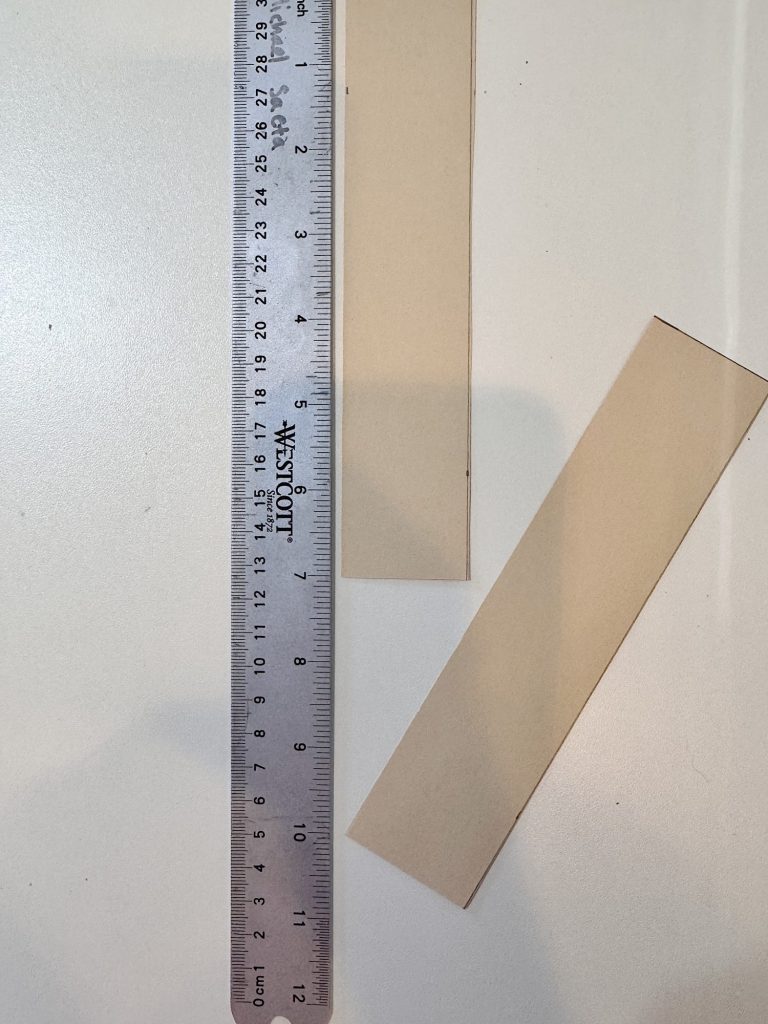 A metal ruler with centimeter markings is placed next to two rectangular strips of light brown cardboard on a white surface.