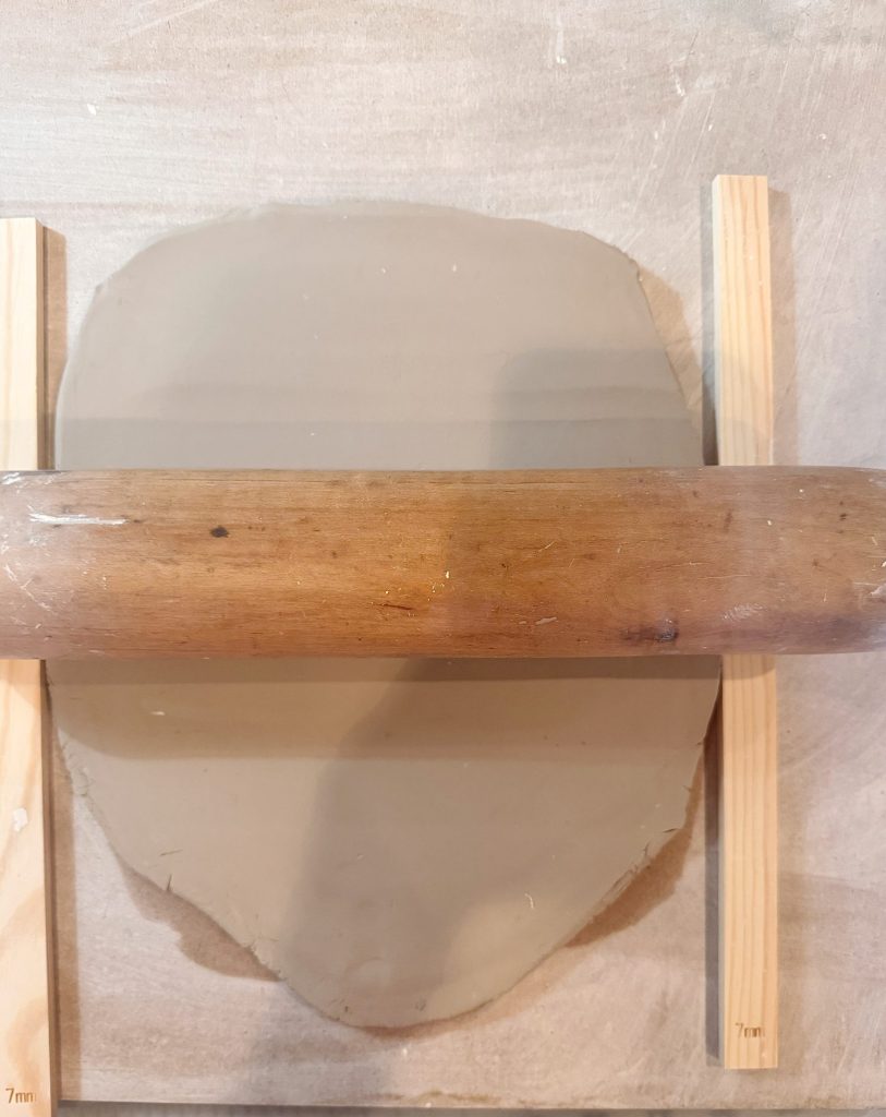 A rolling pin flattens a slab of clay between two wooden slats on a light-colored surface.