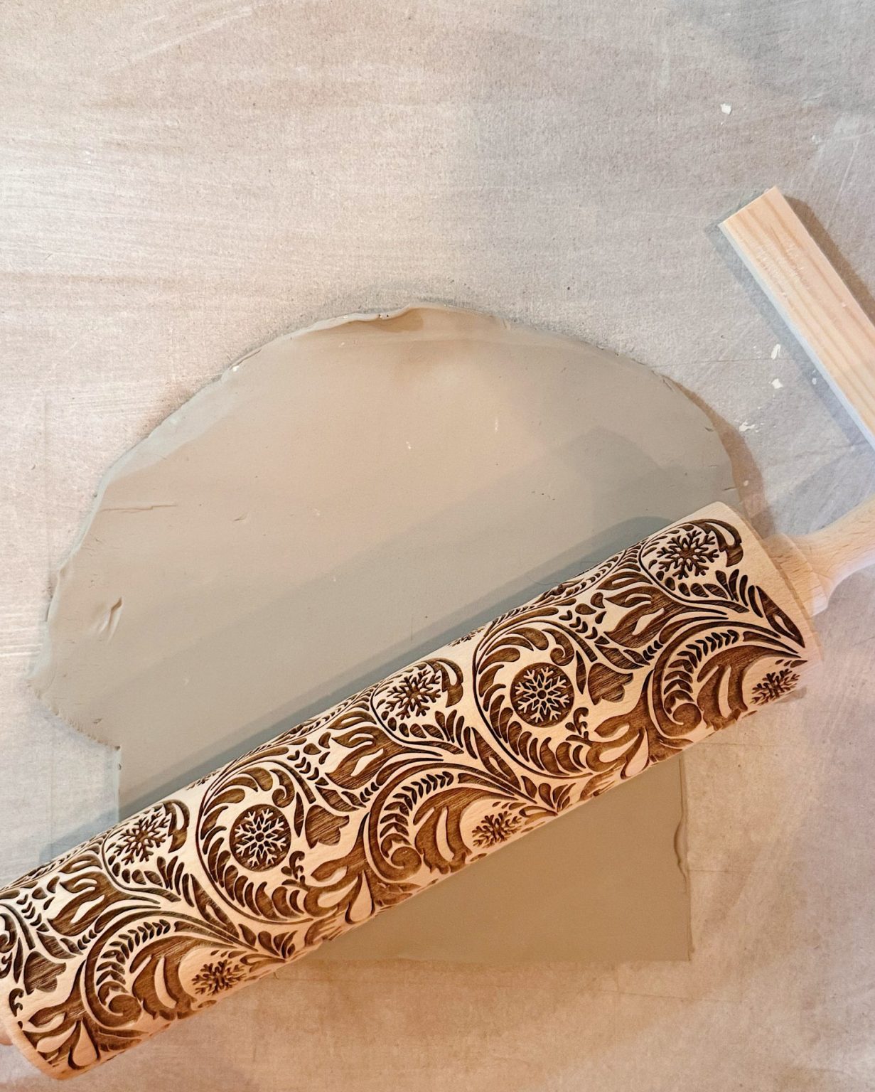 A patterned rolling pin rests on a sheet of clay, leaving decorative floral imprints on the surface.