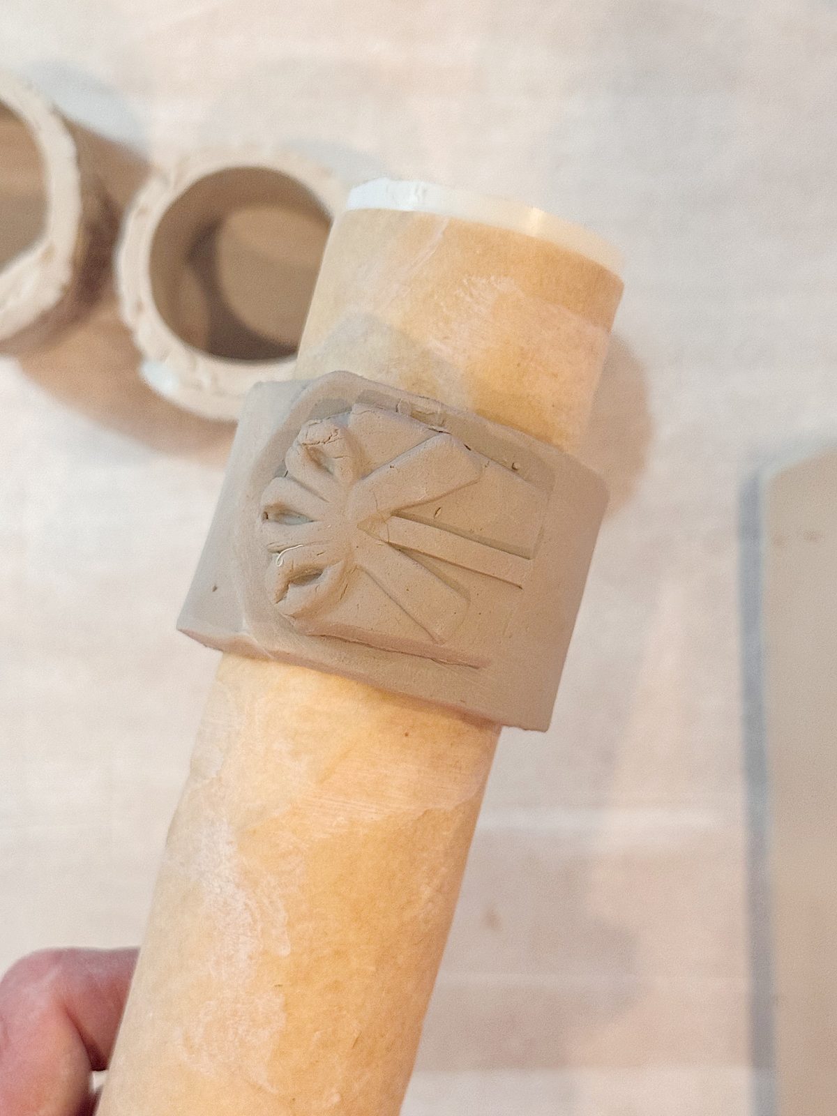 A hand holds a cylindrical object wrapped with a piece of clay that has a sunburst design carved into it. Two circular clay pieces are in the background.