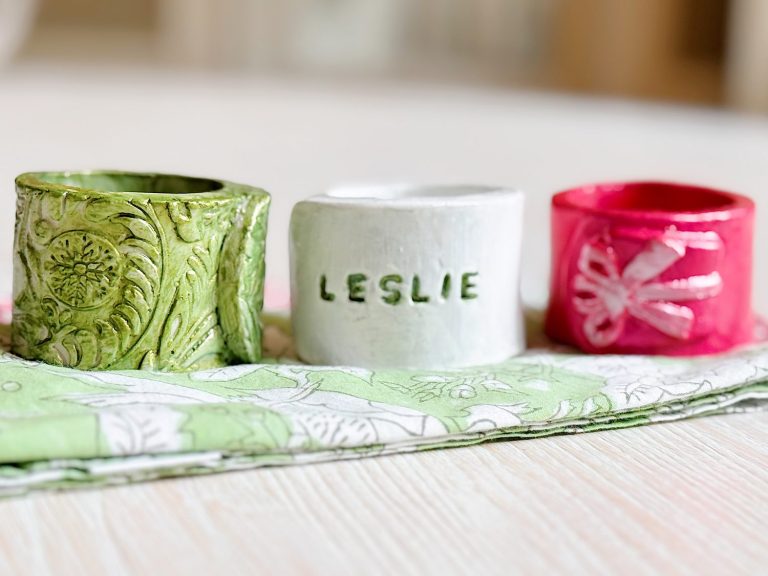 Three handmade napkin rings—one green with a floral pattern, one white with "LESLIE" embossed, and one pink with a dragonfly—rest on folded patterned napkins.