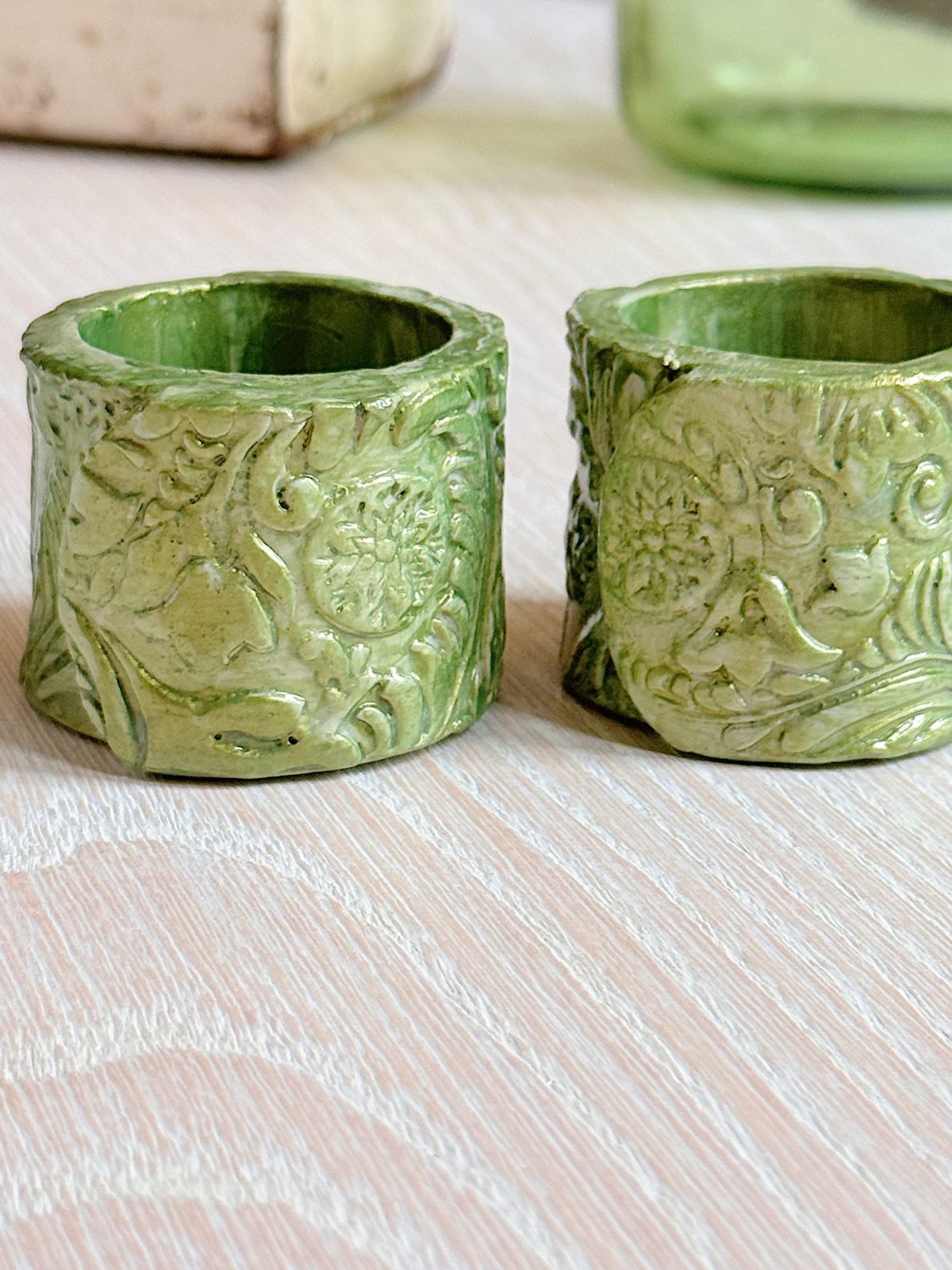 Two green ceramic napkin rings with intricate floral and leaf patterns sit side by side on a light wooden surface.