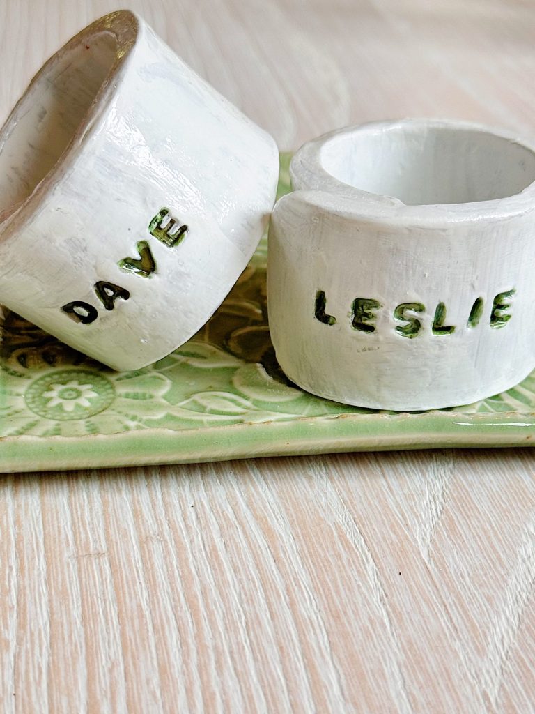 Two white ceramic napkin rings labeled "DAVE" and "LESLIE" sit on a green patterned tray atop a light wooden surface.