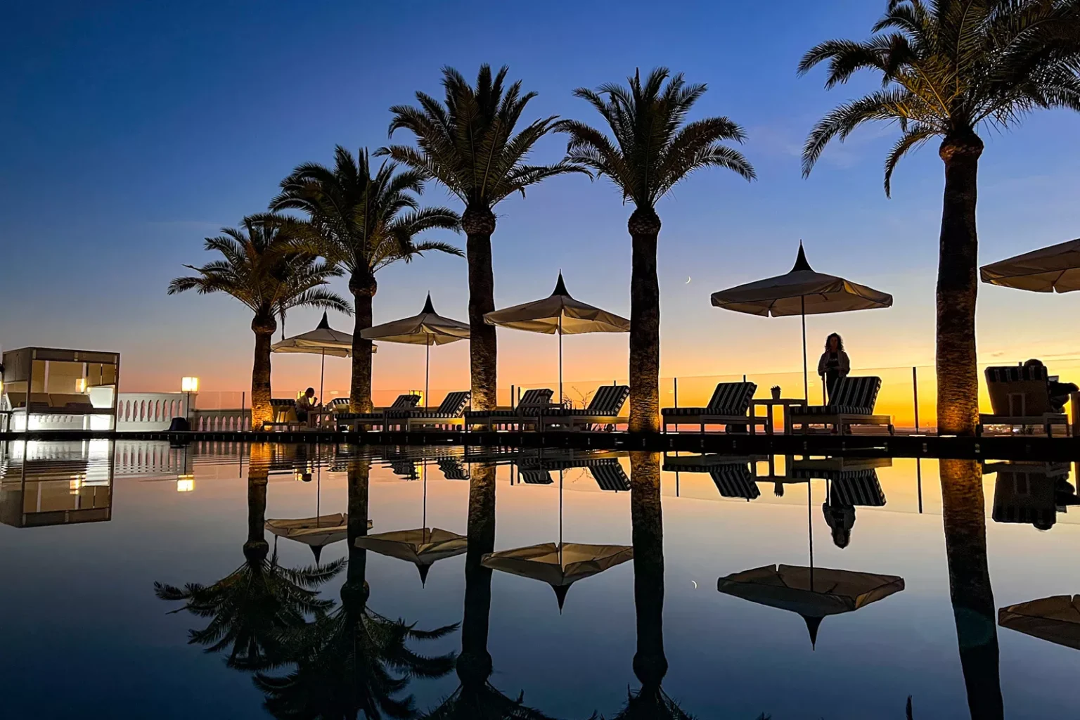 Rows of palm trees and lounge chairs reflected in a calm pool at sunset, with umbrellas and a few people silhouetted against the sky.