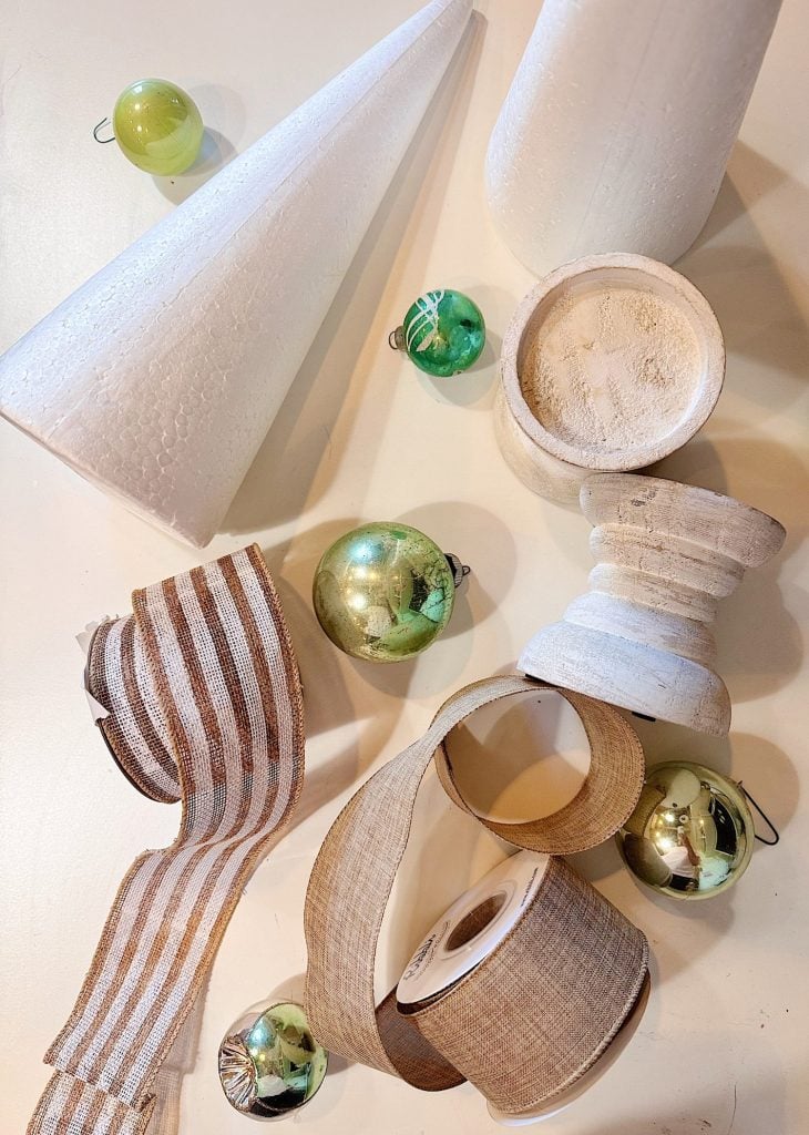 Assorted Christmas craft supplies including green ornaments, white Styrofoam cones, a wooden base, and rolls of ribbon arranged on a light surface.