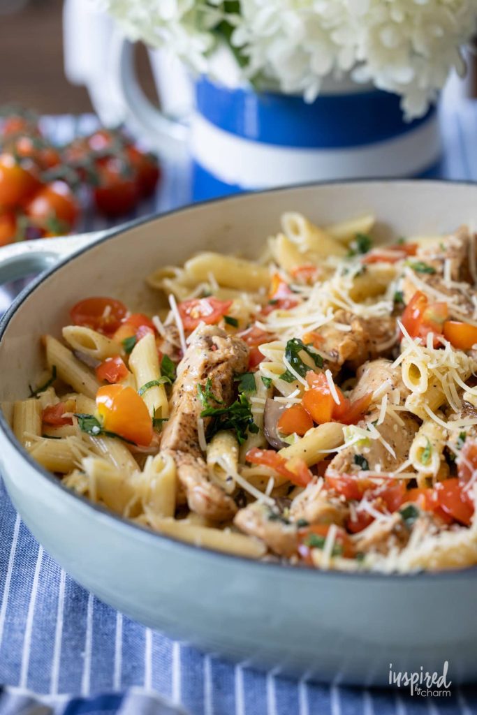 A skillet filled with penne pasta, grilled chicken, chopped tomatoes, fresh herbs, and grated cheese, with a blue-striped pitcher and flowers in the background.
