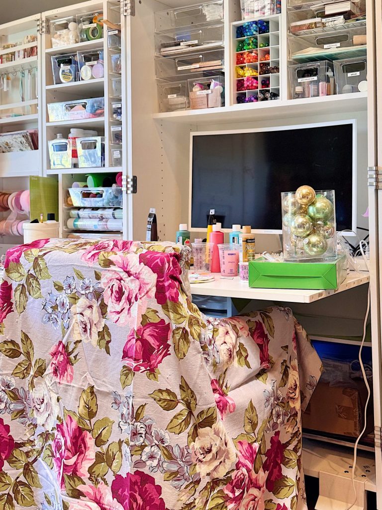 A craft room features shelves of organized supplies, a desk with craft materials, a computer monitor, and a chair covered with a floral-patterned cloth.