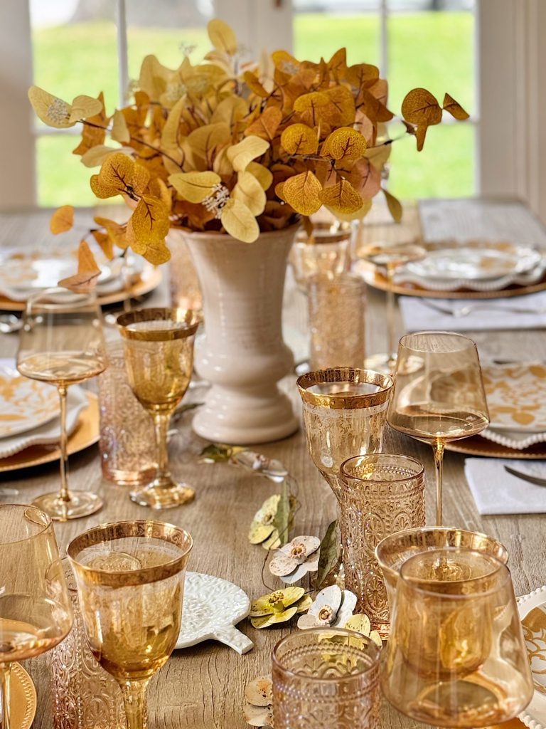 A wooden dining table set with gold-toned glassware, plates, and a white vase filled with yellow autumn leaves as a centerpiece.