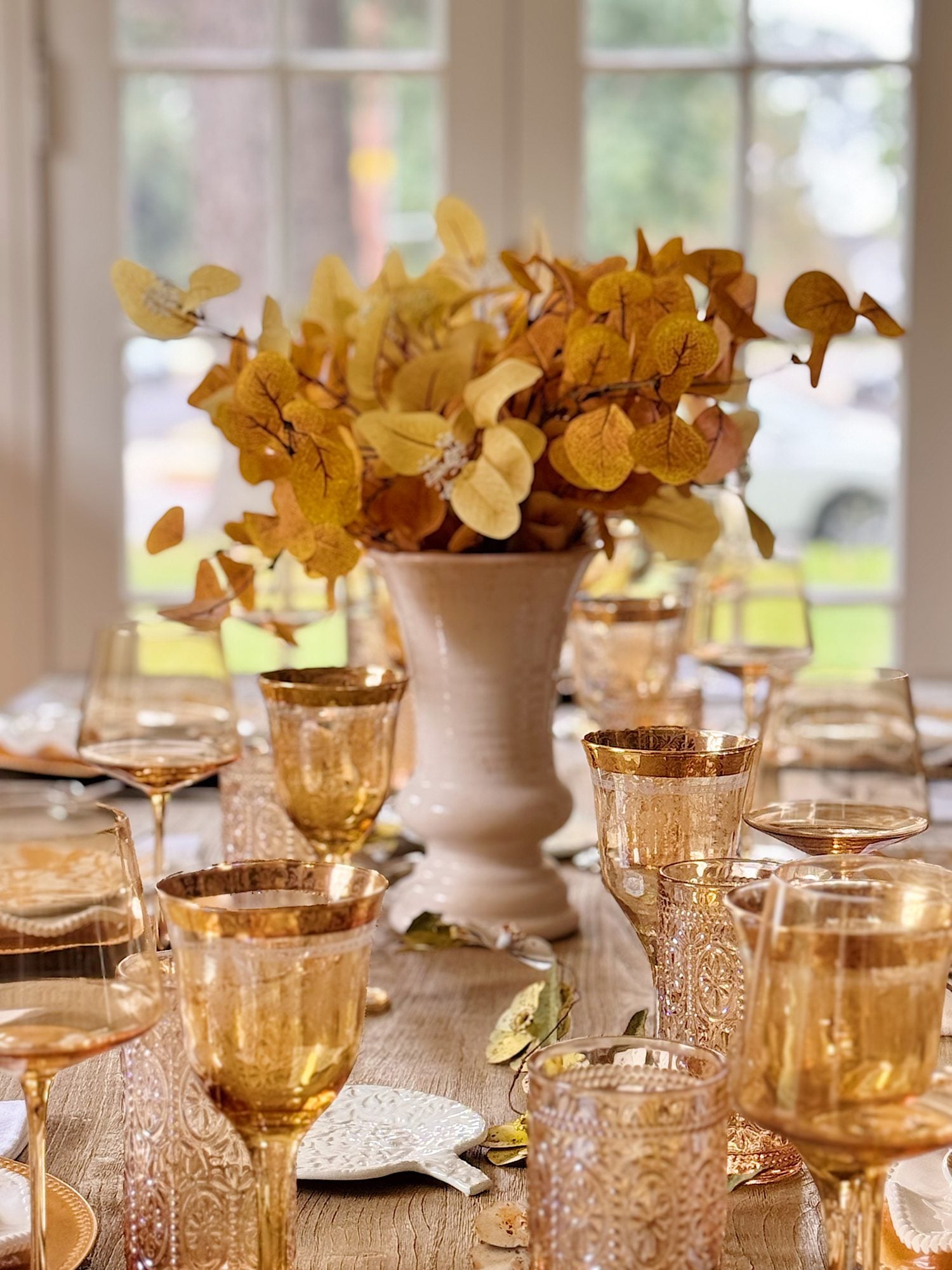 A dining table set with gold-rimmed glassware and a white vase holding yellow autumn leaves, with natural light coming through windows in the background.