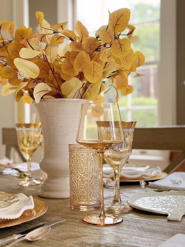 A dining table set with gold-rimmed glassware, white plates, gold flatware, and a large vase of yellow leaves as a centerpiece, with sunlight coming through the window.