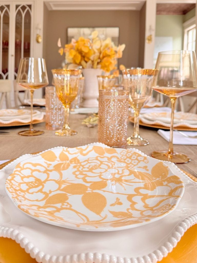 A dining table set with yellow and white floral plates, gold-rimmed glasses, and a centerpiece of yellow flowers in a white vase.