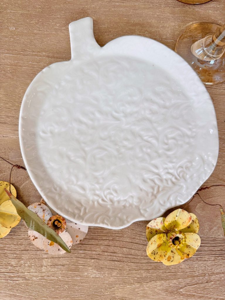 A white ceramic plate shaped like a pumpkin with a textured floral pattern sits on a wooden surface, surrounded by decorative yellow pumpkins and a wine glass.