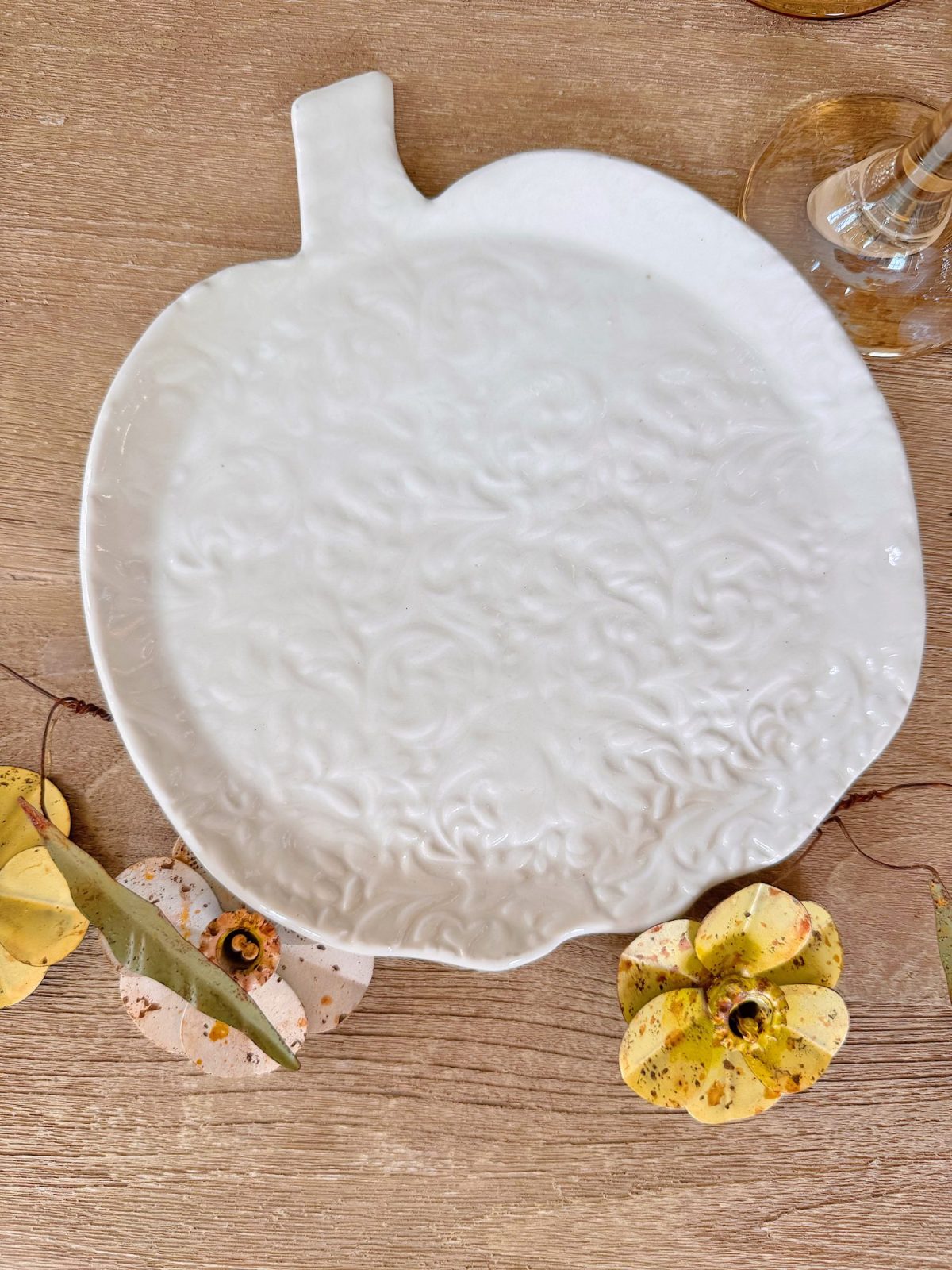A white ceramic plate shaped like a pumpkin with a textured floral pattern sits on a wooden surface, surrounded by decorative yellow pumpkins and a wine glass.