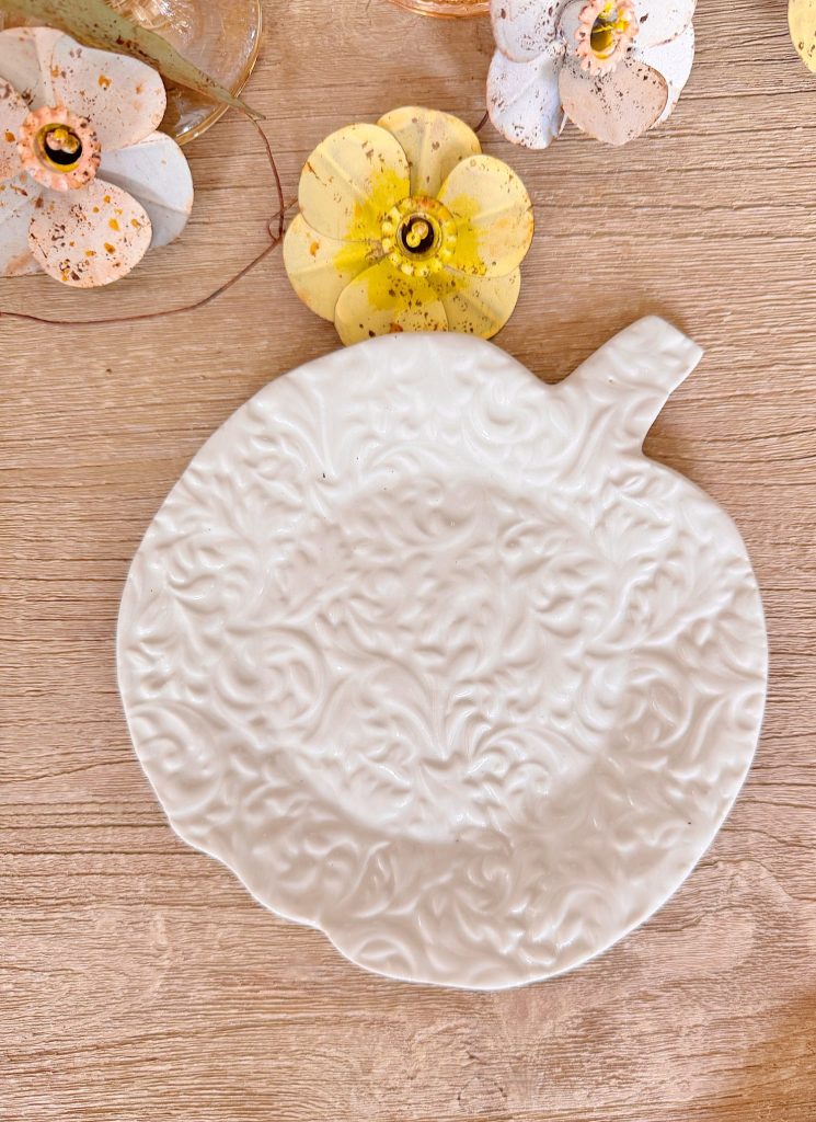 A white ceramic plate shaped like a pumpkin with a textured pattern, placed on a wooden surface with yellow decorative flowers nearby.