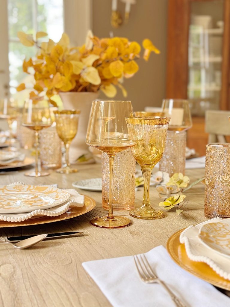 A dining table set with gold-toned glassware, white plates with patterned napkins, silverware, and a centerpiece of yellow foliage in a white vase.