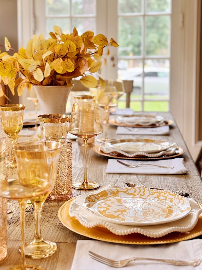 A dining table set with yellow and white patterned plates, gold-rimmed glasses, white napkins, and a vase of yellow leaves, in front of large windows.