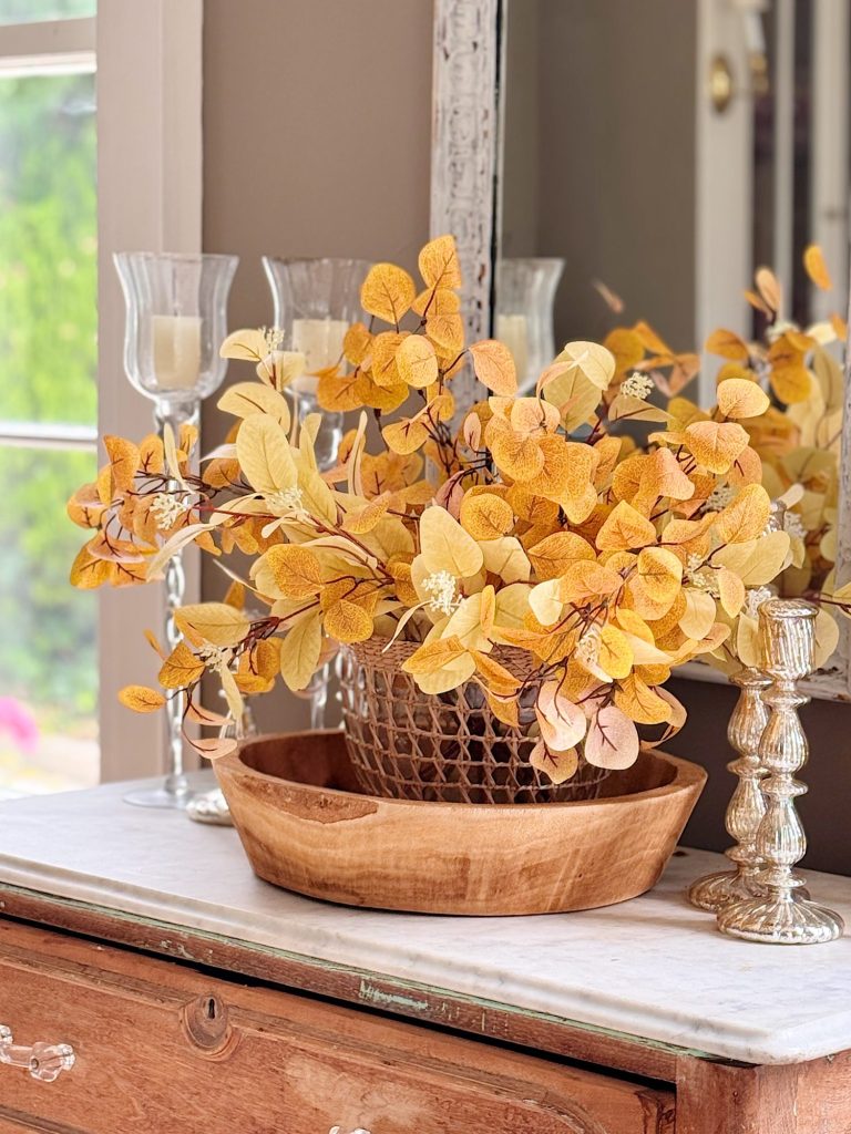 A wooden bowl holds a basket filled with yellow-orange artificial leaves, displayed on a marble-topped dresser beside two glass candle holders.
