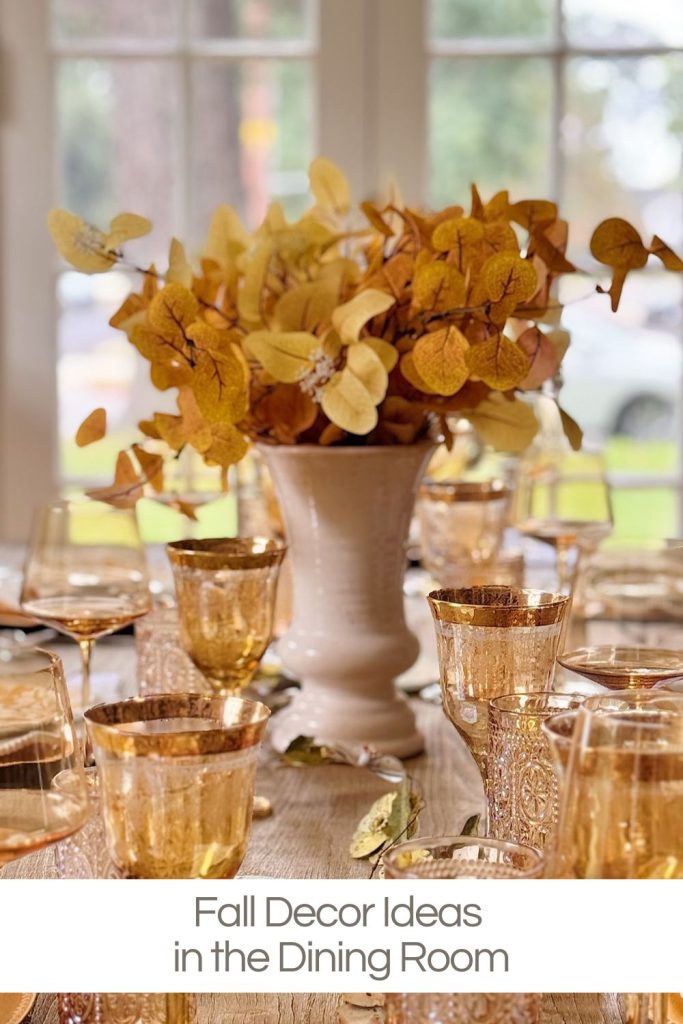 A dining table set with amber glassware and a white vase holding yellow fall foliage, placed in front of large windows. Text reads: "Fall Decor Ideas in the Dining Room.