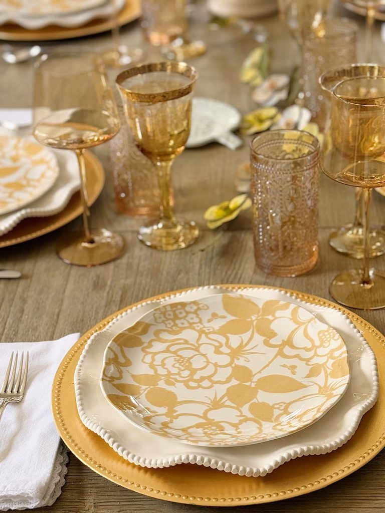 A close-up of a dining table set with gold and white floral-patterned plates, gold chargers, glassware, and white napkins on a wooden surface.