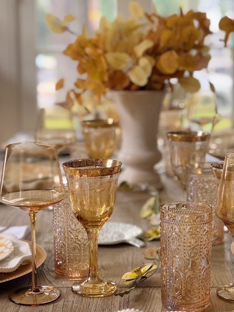 A wooden table is set with amber-tinted glassware, patterned tumblers, wine glasses, plates, and a white vase with yellow foliage as a centerpiece.