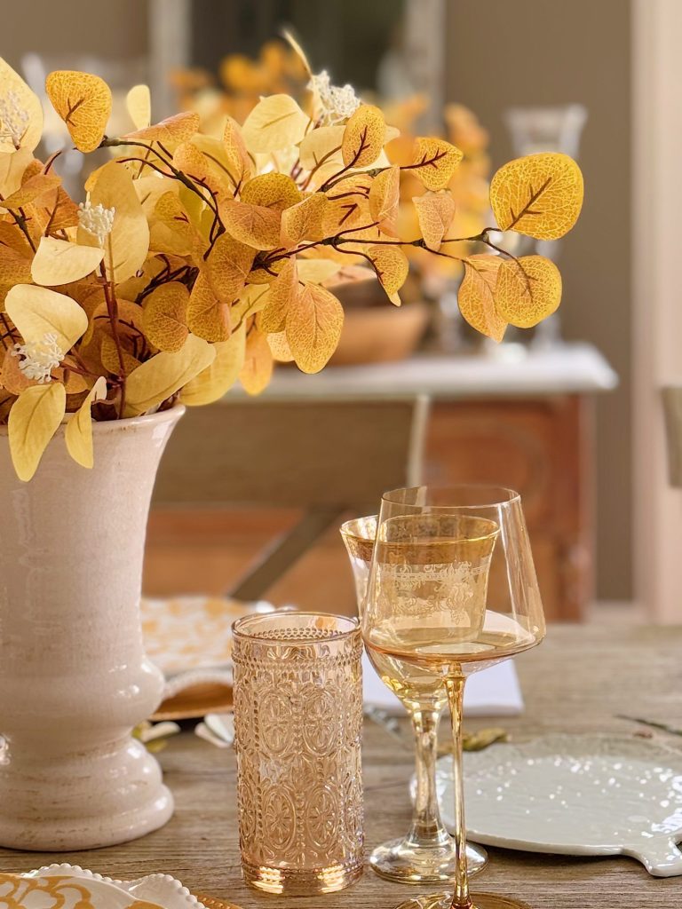 A beige vase with yellow autumn leaves sits on a wooden table set with ornate glassware and a white plate.