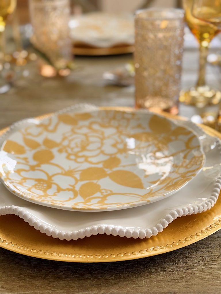A close-up of a decorative table setting with a yellow floral-patterned plate on a white beaded plate, placed on a gold charger, surrounded by textured glasses.