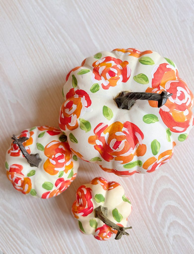 Three white pumpkins decorated with hand-painted red, orange, and green floral patterns are arranged on a light wooden surface.