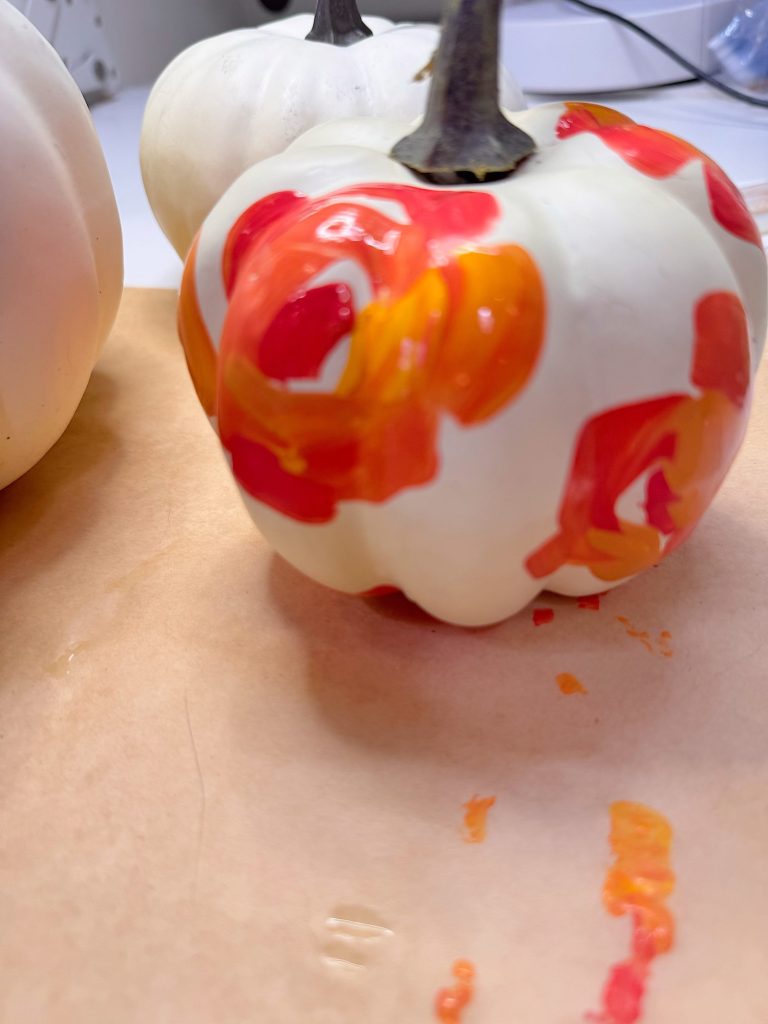 A white pumpkin with orange and red paint marks sits on brown paper, with paint smudges visible nearby and other white pumpkins in the background.
