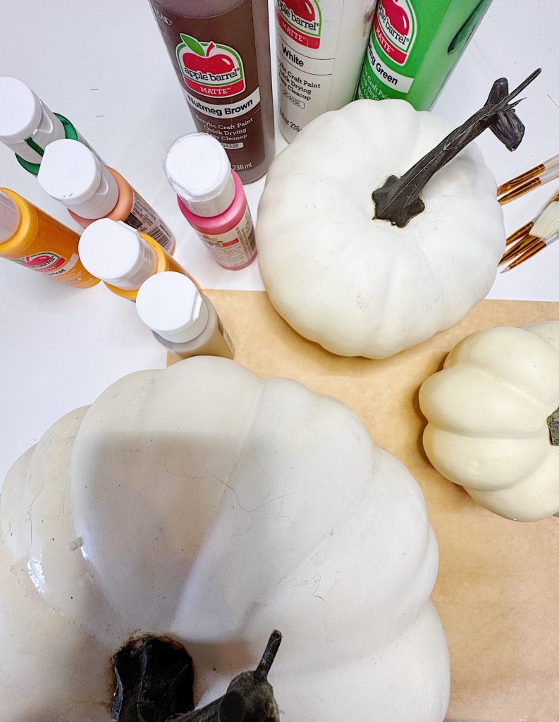 Three white pumpkins, several bottles of acrylic paint in various colors, paintbrushes, and a sheet of brown paper are arranged on a white surface.