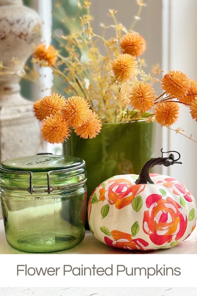 A white pumpkin painted with colorful flower designs sits next to a green glass jar and a vase of orange dried flowers.