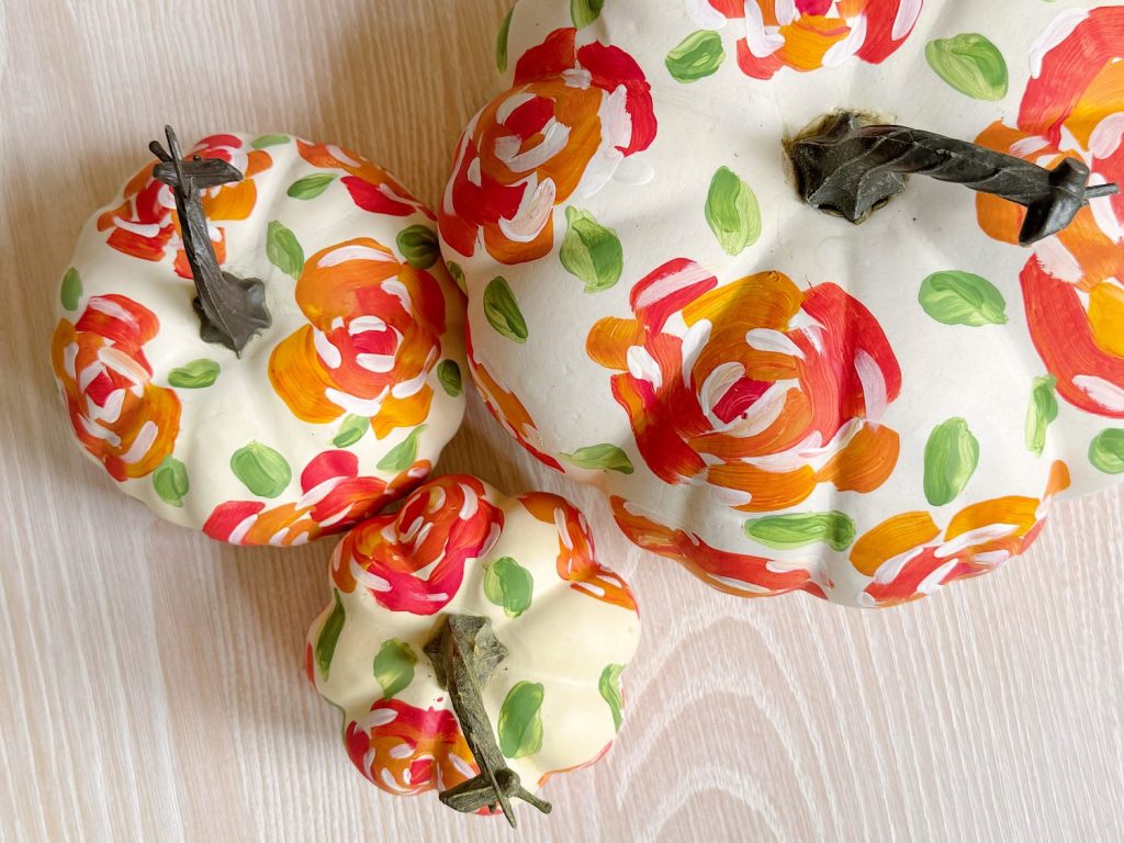 Three white pumpkins with hand-painted red, orange, and pink floral designs and green leaves, arranged on a light wood surface.
