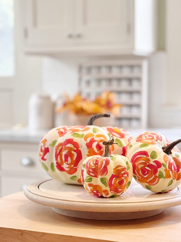 Three white pumpkins with hand-painted red and orange floral designs sit on a round wooden tray in a bright, modern kitchen.