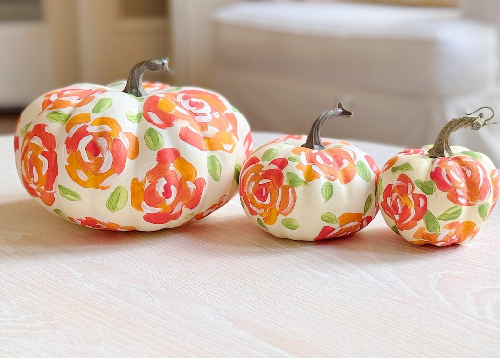 Three white pumpkins of varying sizes are decorated with hand-painted red, orange, and green floral patterns, displayed on a light-colored surface.