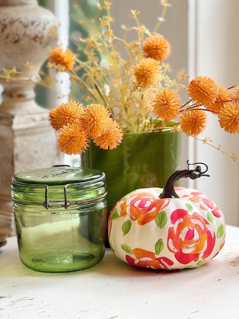 A green glass jar, a green vase with orange dried flowers, and a white pumpkin painted with colorful floral patterns sit on a white surface.