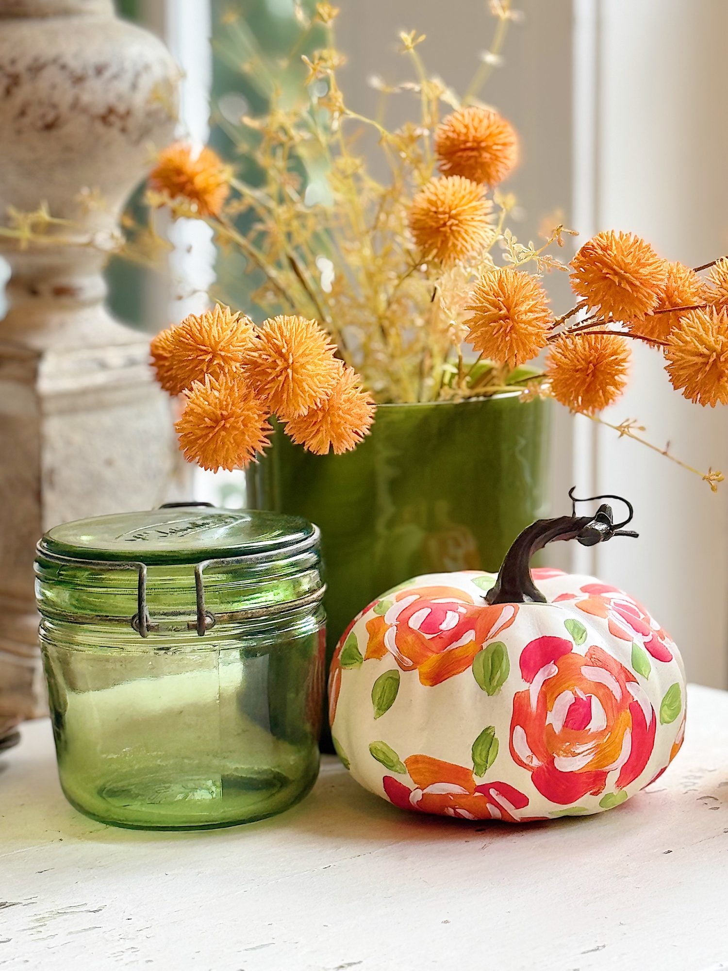 A green glass jar, a green vase with orange dried flowers, and a white pumpkin painted with colorful floral patterns sit on a white surface.