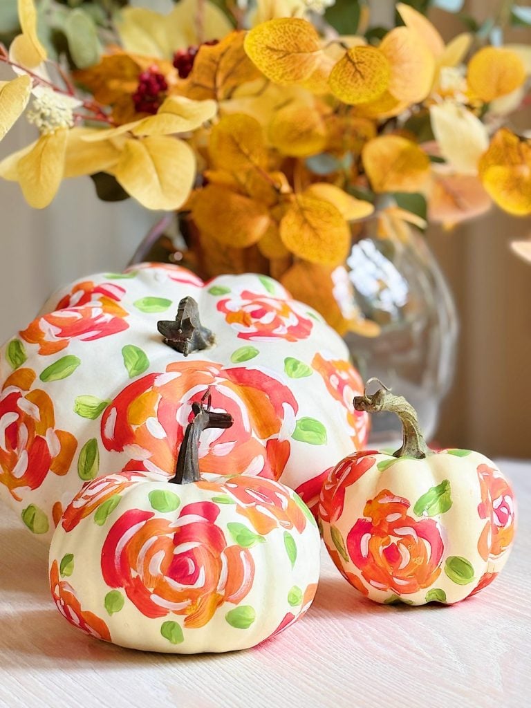 Three white pumpkins are decorated with hand-painted orange, green, and pink floral designs, set in front of a vase with yellow autumn leaves.