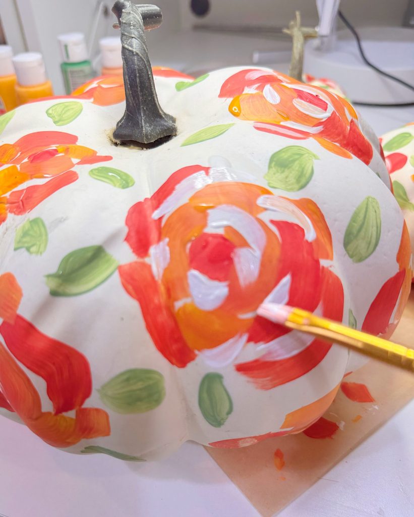 A hand uses a paintbrush to paint orange, red, and white flower patterns on a white pumpkin; green leaves are also visible around the flowers.