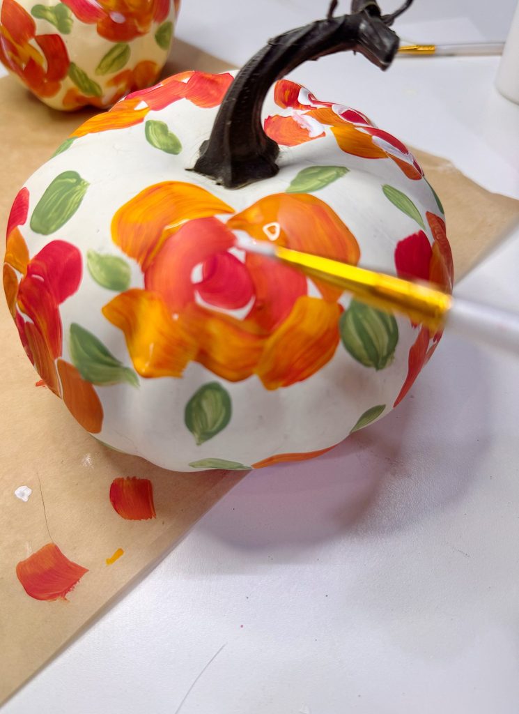 A hand paints orange and red flowers with green leaves on a white decorative pumpkin using a small paintbrush.