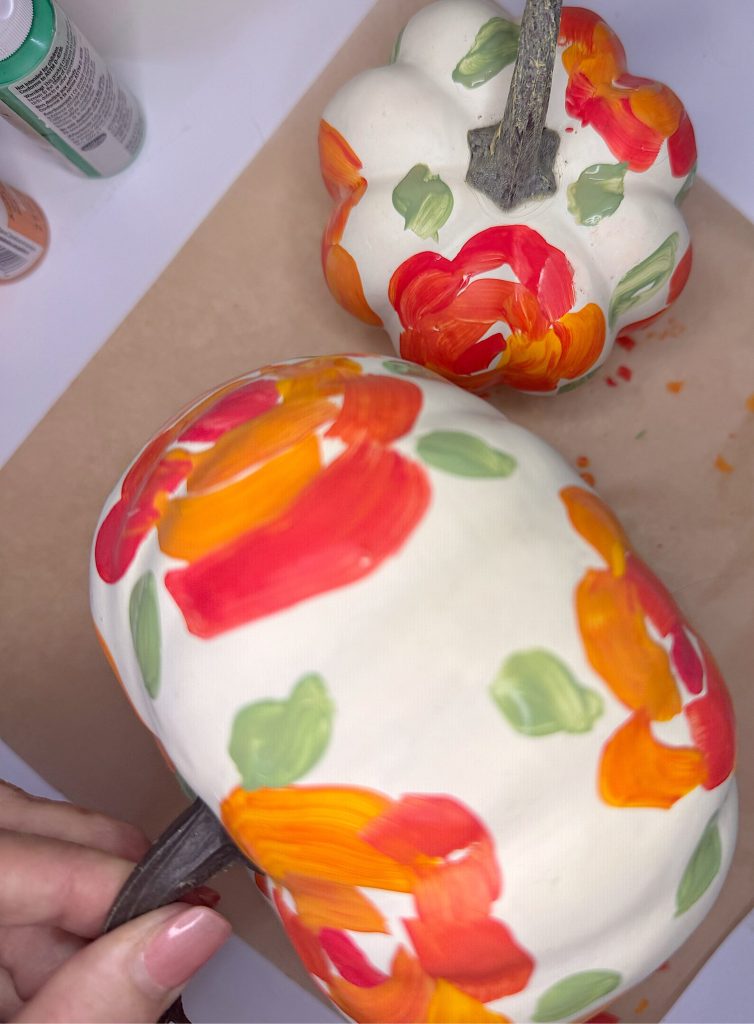 Two white pumpkins are decorated with hand-painted orange, red, and yellow flowers with green leaves. A hand holds the stem of one pumpkin. Paint bottles are visible in the background.