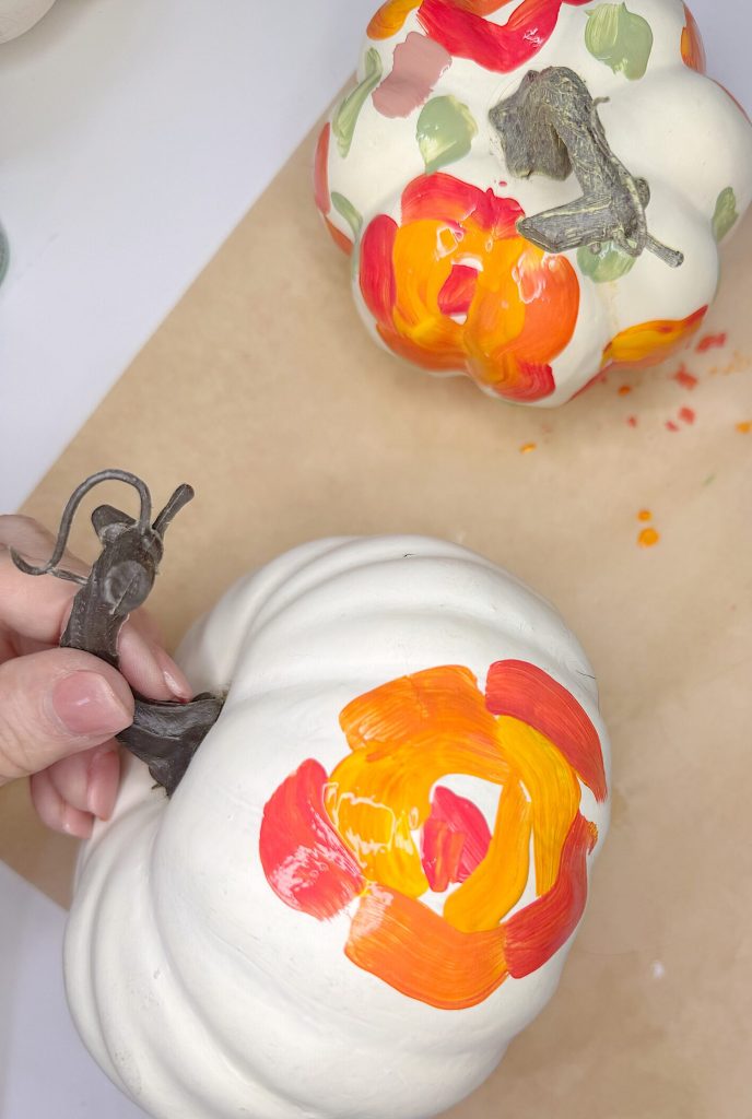A hand holding a white pumpkin with orange and red painted designs next to another similarly painted pumpkin on a tan surface.