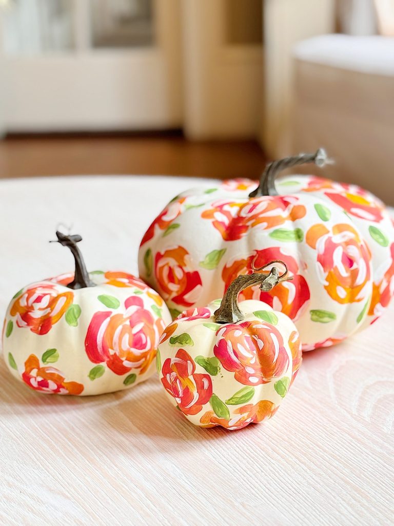 Three white pumpkins with painted red, orange, and green floral designs are displayed on a light-colored wooden surface indoors.