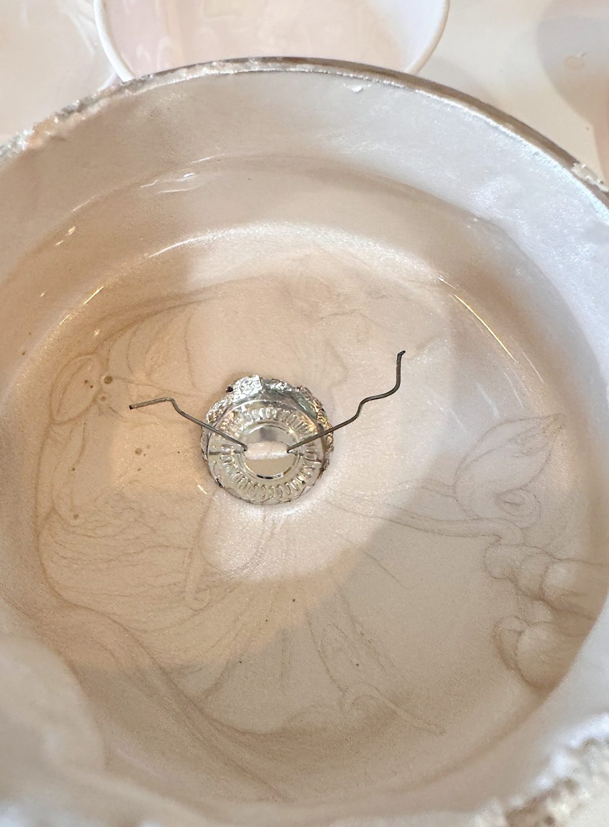 A close-up of a round ceramic bowl with a transparent liquid inside, featuring a metal bottle cap and a bent wire partially submerged at the bottom.