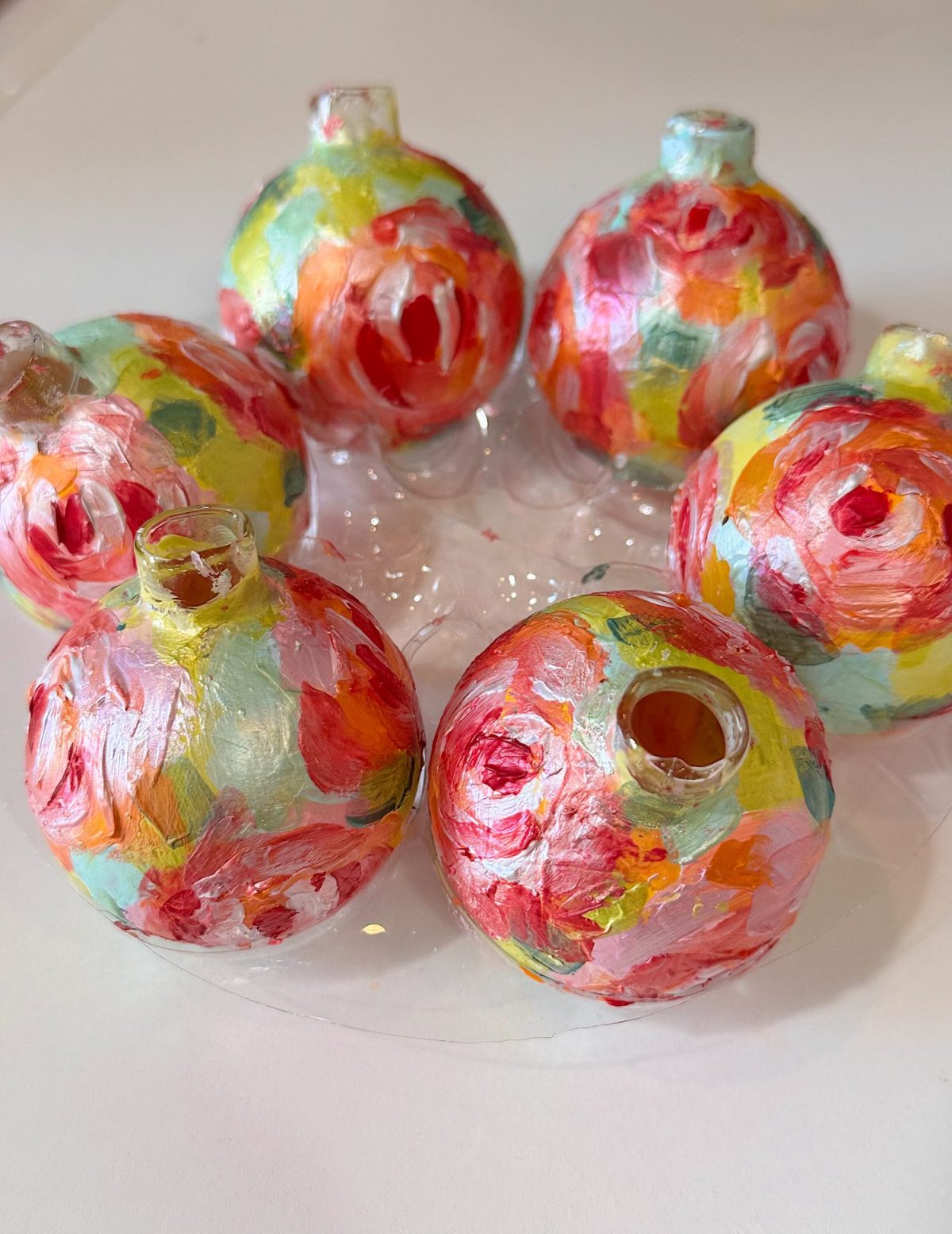 Six round glass ornaments painted with colorful, abstract brushstrokes in red, pink, yellow, and green rest on a clear plastic tray.
