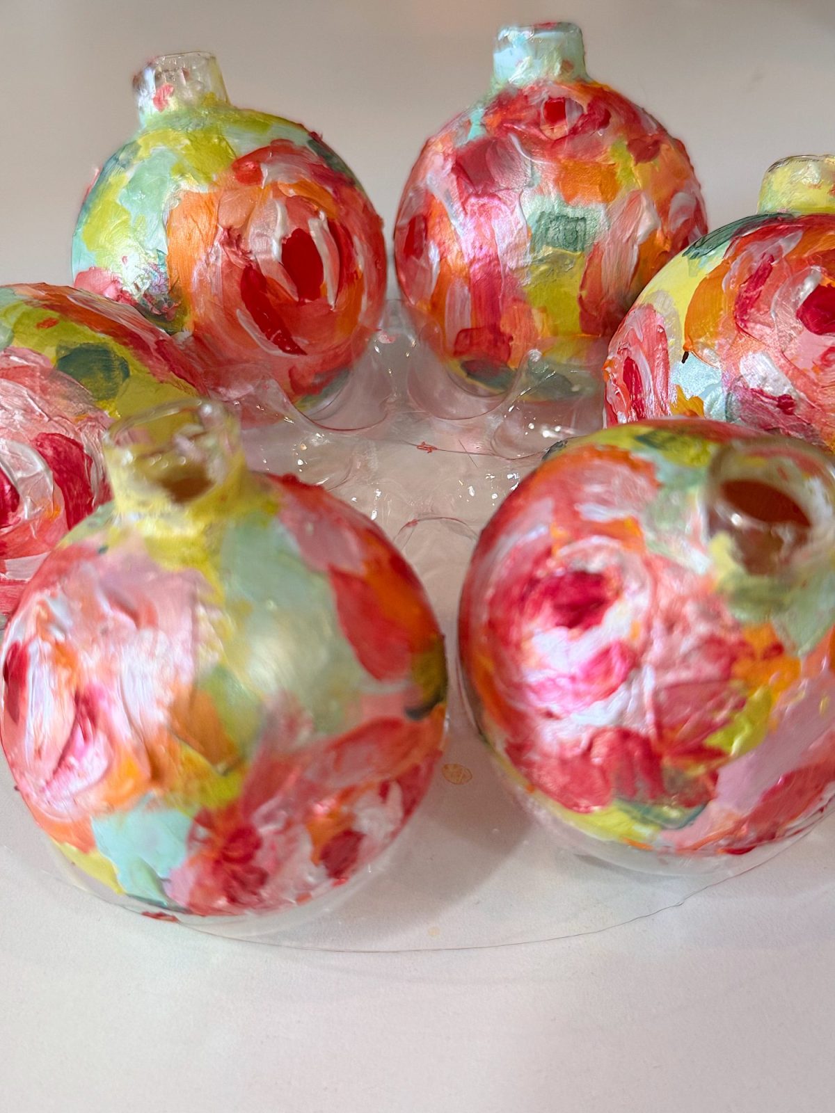 Six glass ornaments painted with colorful, abstract floral designs are arranged in a circle on a clear plastic tray.