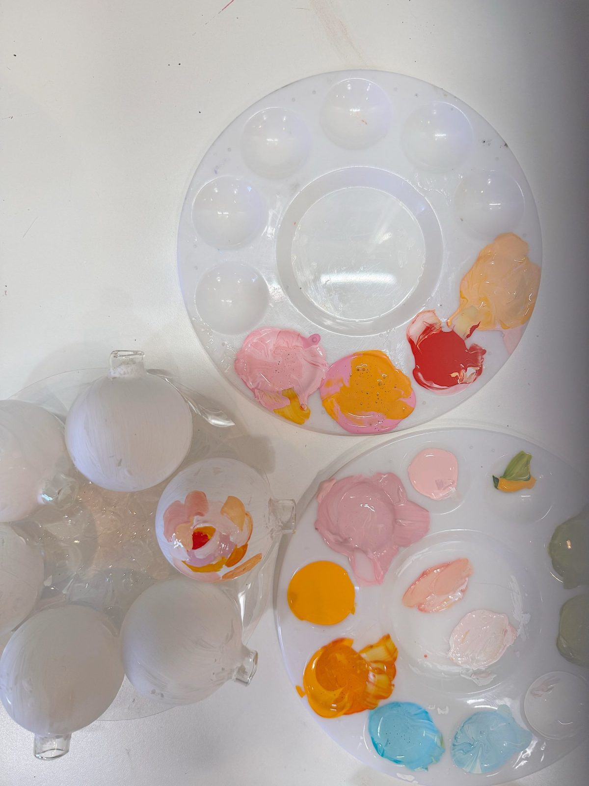 Two paint palettes with various pastel paint colors, and several white round objects, possibly ornaments, being decorated on a white surface.