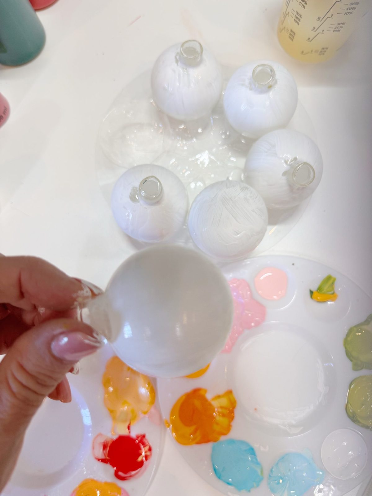 A hand holds a white ornament above a table with more white ornaments, paint palettes, and various paint colors.