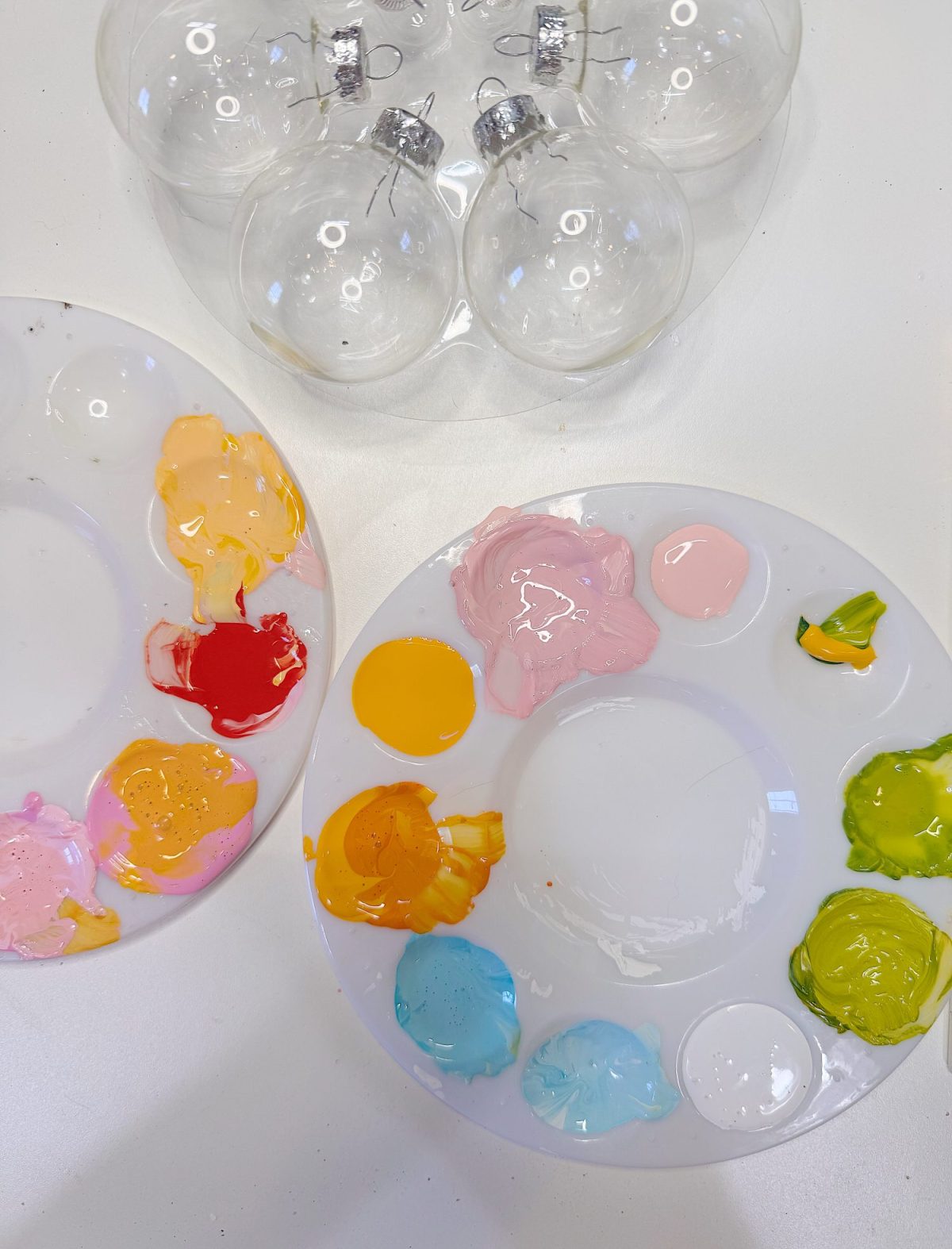 Two paint palettes with various colorful paints are placed next to four clear glass ornaments on a white surface.