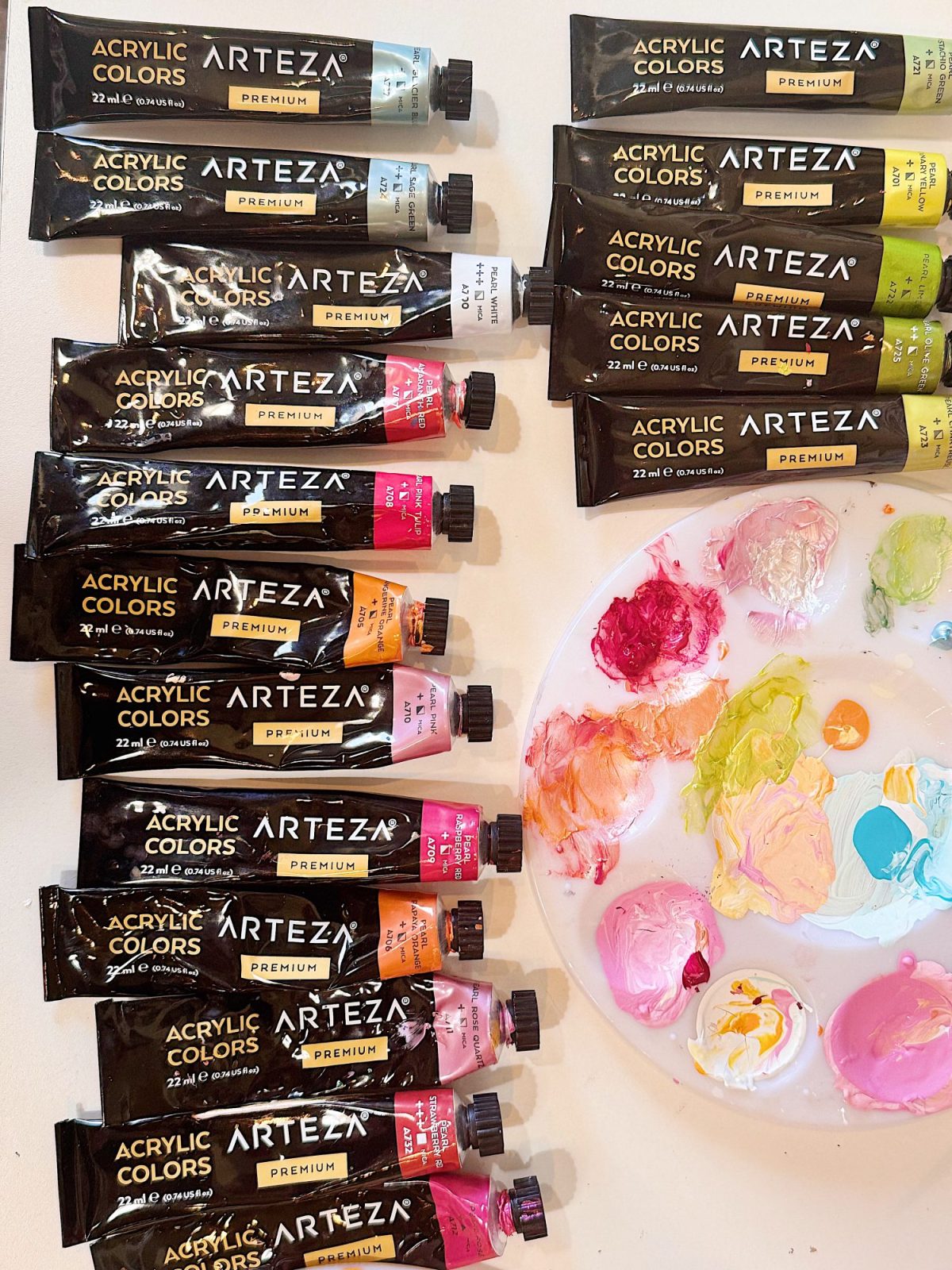 Tubes of Arteza acrylic paint are arranged next to a palette with mixed pastel colors on a white surface.