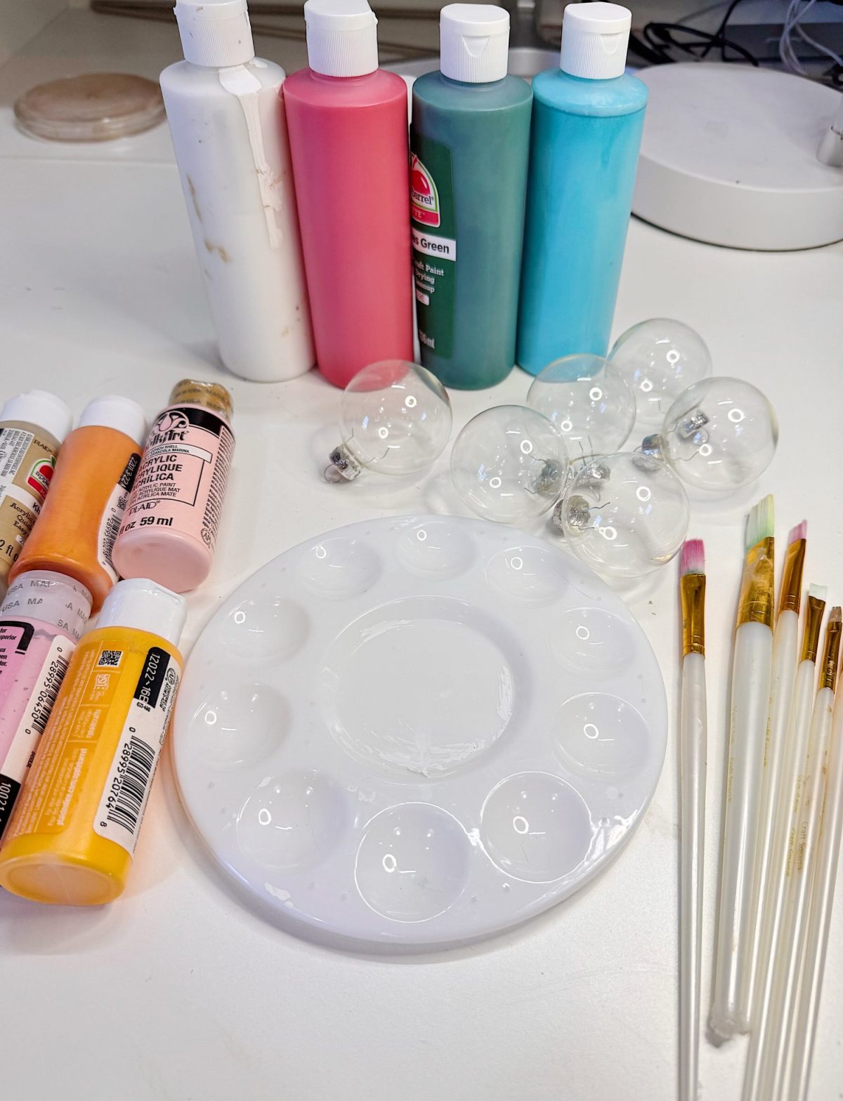Acrylic paint bottles, paintbrushes, empty clear ornaments, and a plastic paint palette arranged on a white table.