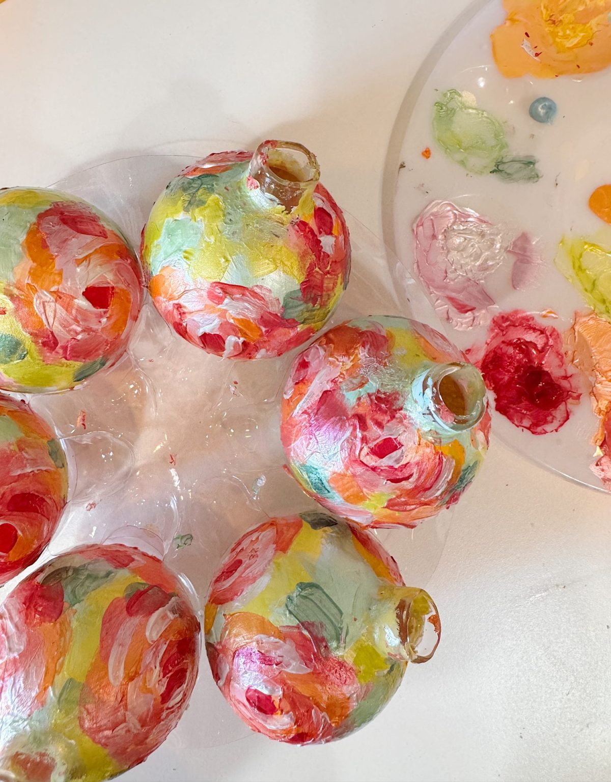 Five glass ornaments are painted with colorful, abstract brushstrokes, and a paint palette with mixed colors sits nearby on a white surface.